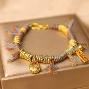 Tibetan lucky fortune bracelet with gold coil accents and red detail charm on beige jewelry box display, showing frayed rope tassel ends and lobster clasp