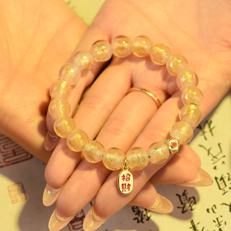 Tibetan fortune bracelet light gold glass beads with zhao cai charm resting on palm over calligraphy paper