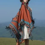 Tibetan orange geometric tassel shawl worn outdoors in mountain landscape, terracotta base with ethnic tribal patterns and fringed hem