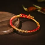 Tibetan gold and red braided protection bracelet with dragon scale knot, three-quarter view on wooden coaster with dried botanical elements
