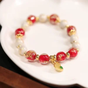 Red and white Tibetan god of wealth gourd bracelet with gold leaf charm pendant displayed on white porcelain, top-down flat lay showing liuli glass and incense ash beads