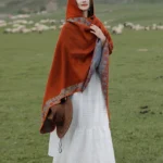 Tibetan hooded floral wrap shawl in burnt orange worn outdoors with grassland and sheep flock background, front three-quarter view