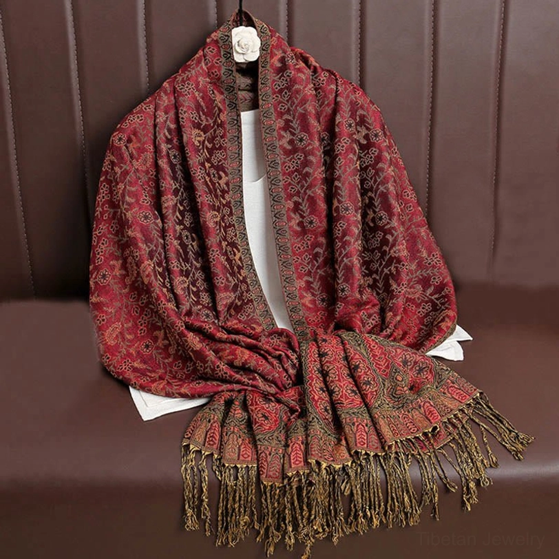 Burgundy red Tibetan tassel shawl with brown trim and all-over paisley jacquard pattern, hung on sofa backrest highlighting decorative bottom fringe
