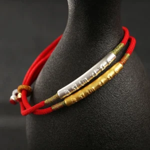 Layered Tibetan red string protection bracelets with dual gold and silver mantra tubes displayed on ceramic stand