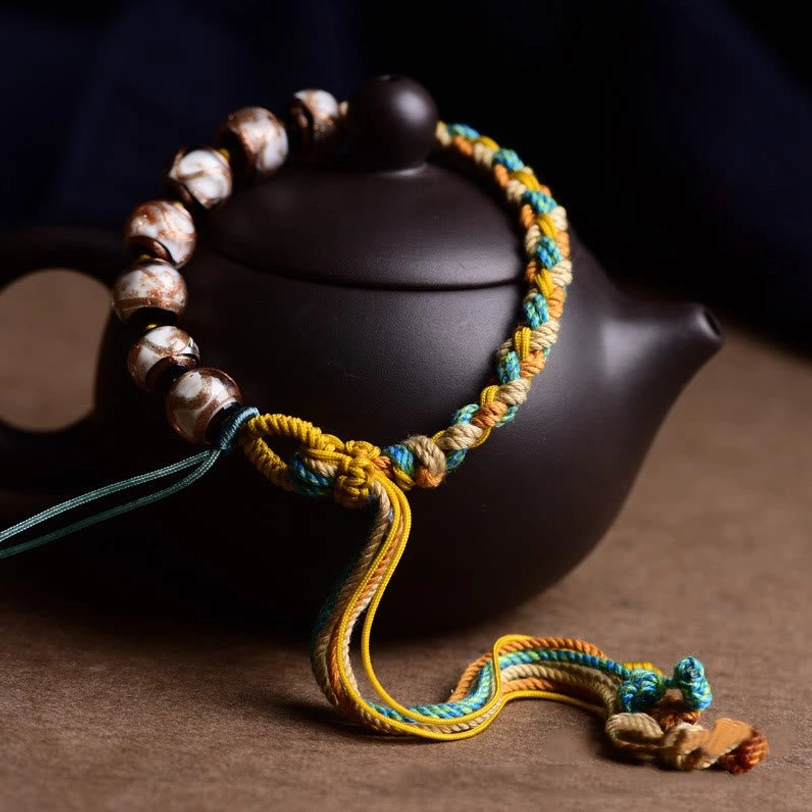 Adjustable Tibetan liuli glass bracelet with yellow blue khaki braided cord and glazed beads, three-quarter angled view on black teapot and brown surface