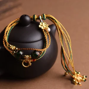 Tibetan five elements liuli glass healing bracelet displayed overhead on black teapot, showing multi-layered braided design with golden green and brown woven cords
