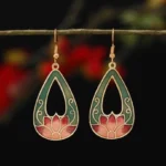 Hollow teardrop lotus enamel drop earrings in dark green and pink with gold tone trim, hung on delicate twig with red floral background