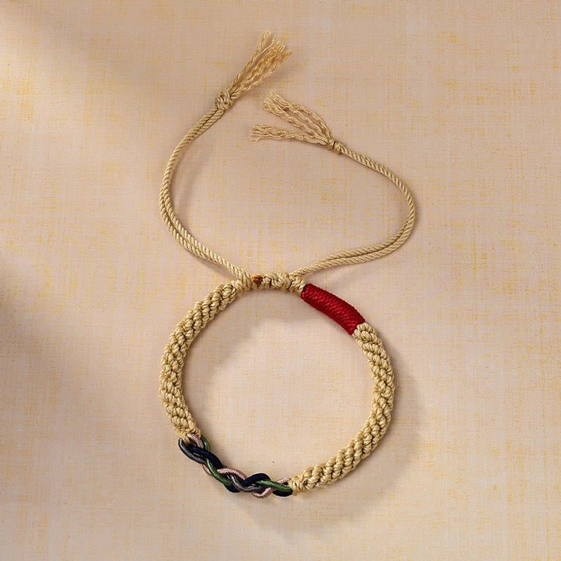 Top-down view of hand-braided Tibetan luck bracelet with golden main rope, red accent knot, and dark twisted decorative knot on tan background