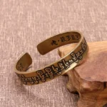 Tibetan Mahāpratisarā Mantra copper brass open cuff bracelet 3/4 view with Sanskrit sutra engravings on linen and wooden display
