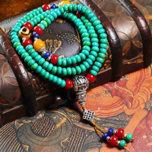 Complete Tibetan mala prayer beads with nine-eye dzi bead arranged on wooden treasure chest and ethnic textile background, showing bright teal beads, red and yellow accent stones, ornate silver guru bead, and traditional tassel