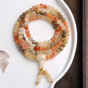 Light-toned natural agate 108-bead mala bracelet arranged on white porcelain dish, featuring creamy white, pale yellow and light reddish polished beads with smooth finish and tassel pendant