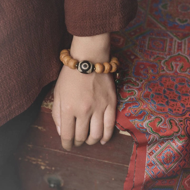 Worn Tibetan bodhi dzi bead mala bracelet on adult wrist with three-eyed dzi focal bead and ethnic patterned carpet background