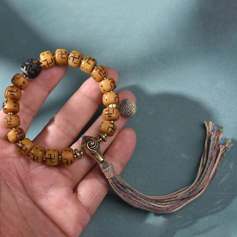 Hand-held view of Tibetan mala bracelet showing carved tan beads, black spacer beads, metal mala counter, silver charm, and multicolor tassel