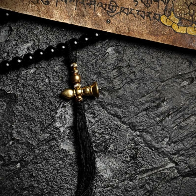 Mala bead tail set close-up showing brass bell-shaped guru bead pendant with vintage brass accents and black onyx on dark stone background