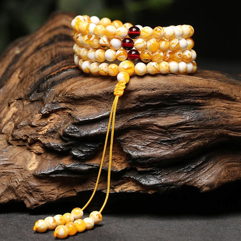 Multi-row Tibetan mala bracelets and long prayer mala with yellow-white beads, dark red spacers, and golden tassel on rough wood surface