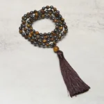 Tiger Eye and Hematite 108 bead mala necklace with brown tassel laid out on light gray background, showing mixed stone beads in warm brown and metallic tones