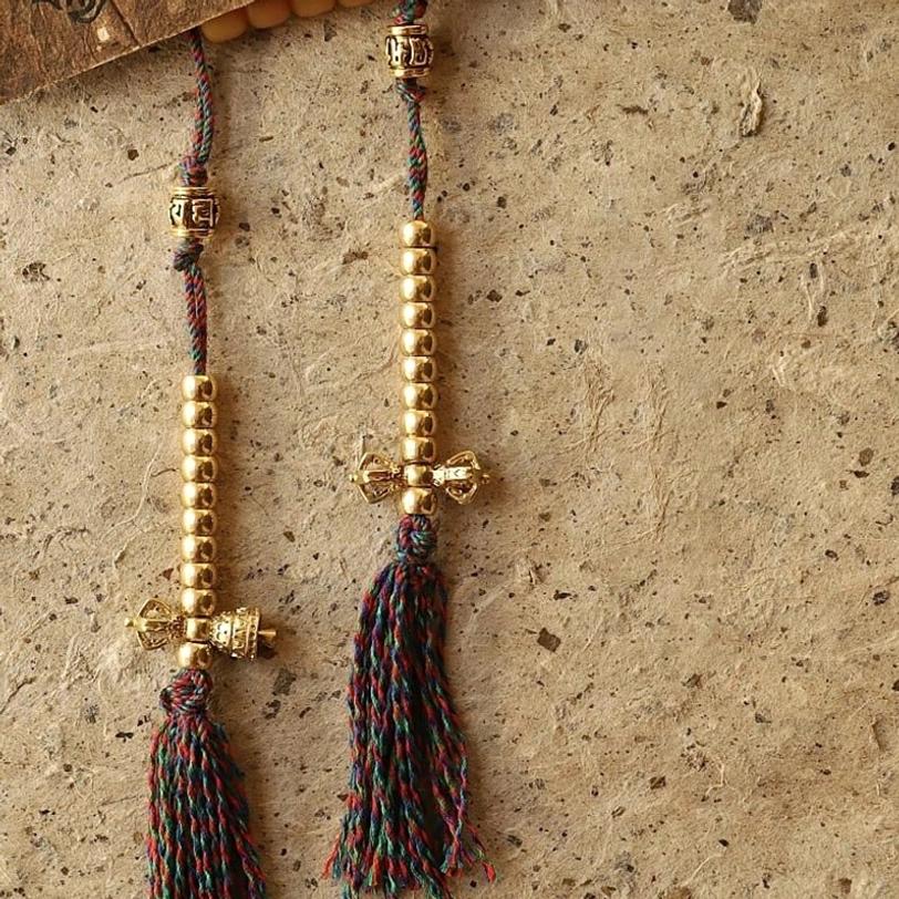 Tibetan mala with vajra counter beads featuring vajra-shaped gold metal charms, multi-color braided cords, and tassel ends on stone wooden surface