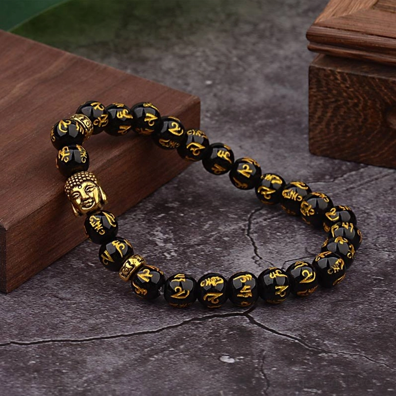 Side angle view of handmade Om Mani Padme Hum bracelet with black glossy beads, gold mantra script, and gold Buddha charm on natural wood and stone