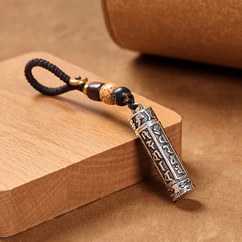 Tibetan mantra keychain three-quarter view showing engraved copper tube pendant with dark brown braided rope and decorative beads