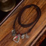 Tibetan Medicine Buddha necklace flat lay on wooden surface, showing braided brown rope cord with silver-toned triangular amulet and red accent beads