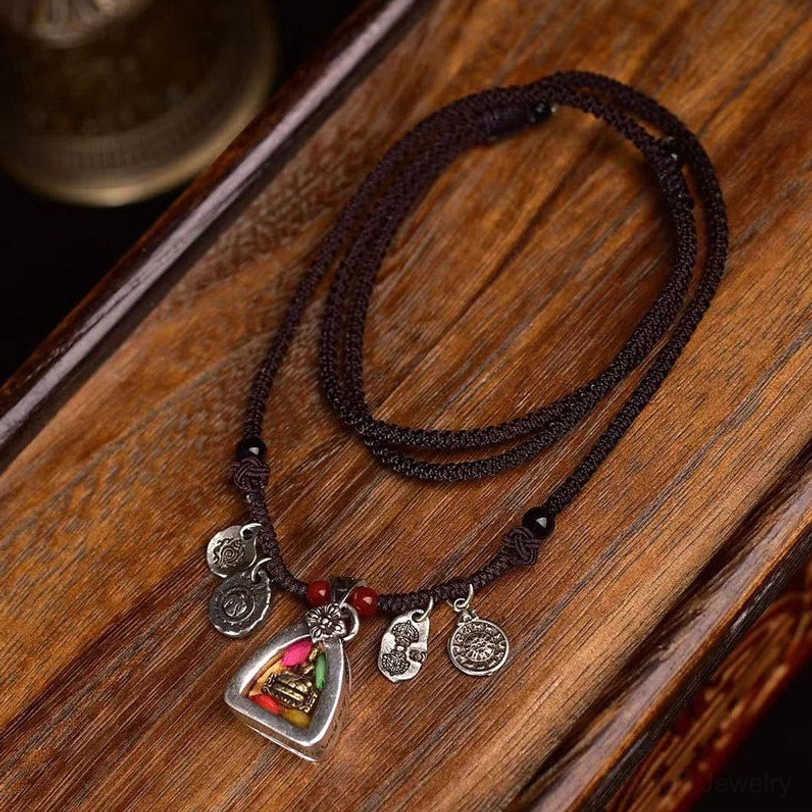 Tibetan Medicine Buddha necklace flat lay on wooden surface, showing braided brown rope cord with silver-toned triangular amulet and red accent beads