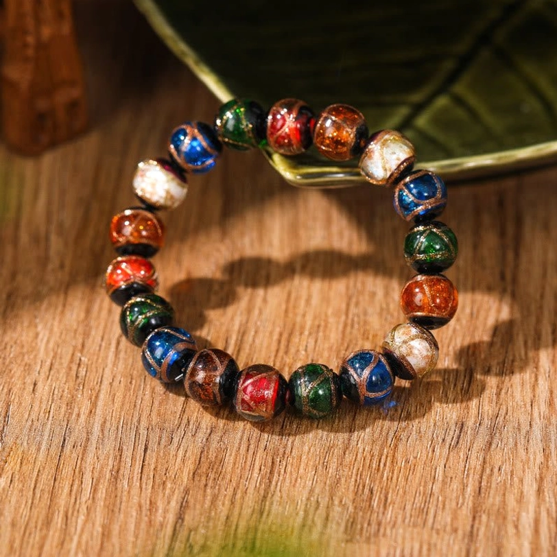 Single mixed-color Tibetan glass beaded bracelet with multicolor glazed beads and gold line details displayed on wooden table with green leaf decoration