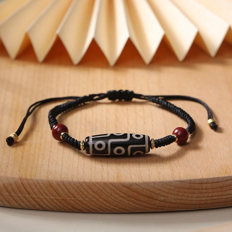 Tibetan nine-eye dzi bead bracelet with traditional design, woven black rope and red beads on vintage wooden table with paper fan