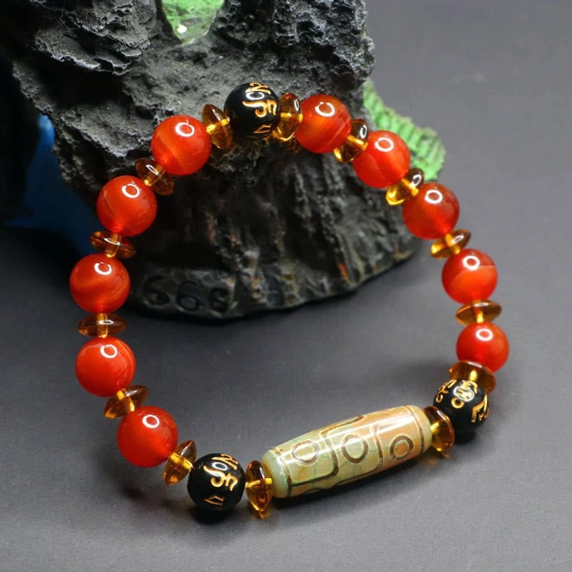Tibetan nine-eye dzi red agate bracelet with vintage dzi bead pendant and black engraved spacer beads, displayed on dark rock ornament