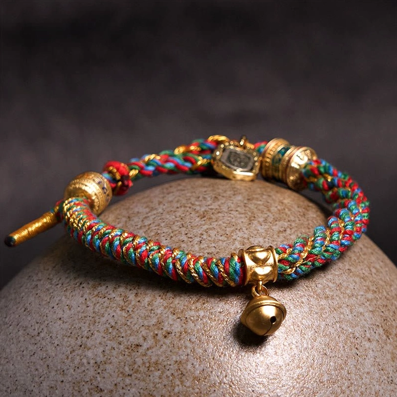 Tibetan mantra bracelet with colorful woven rope and gold fittings displayed on natural stone sphere, showcasing dangling brass bell pendant