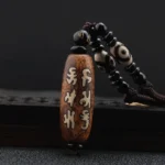 Tibetan Om Mani Padme Hum Dzi bead necklace pendant with hand-painted Sanskrit script on aged brown agate, displayed flat on dark studio background with white beaded cord