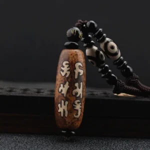 Tibetan Om Mani Padme Hum Dzi bead necklace pendant with hand-painted Sanskrit script on aged brown agate, displayed flat on dark studio background with white beaded cord