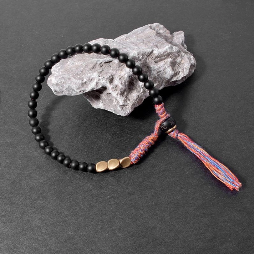 Tibetan black onyx grounding bracelet draped over rough stone with flat gold beads and mixed-thread tassel on dark background