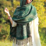 Woman wearing dark teal Tibetan jacquard paisley shawl with light green floral motifs in woodland setting, showcasing three-quarter side view and woven tassel fringe detail