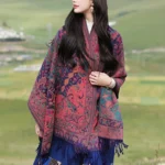Red paisley Tibetan shawl worn by model in outdoor grassland setting, showcasing cashew floral pattern with purple and green tones and decorative side tassels