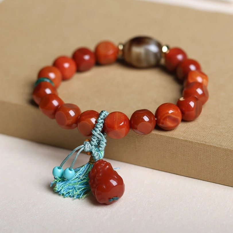 Handcrafted South Red Agate Pixiu Bracelet - Three-Quarter View Showing Faceted Red Agate Beads, Brown Dzi Bead, and Turquoise Tassel on Cardboard Display