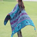 Tibetan geometric floral stripe poncho shawl back view in teal-purple with tassel hem, worn outdoors on grassland