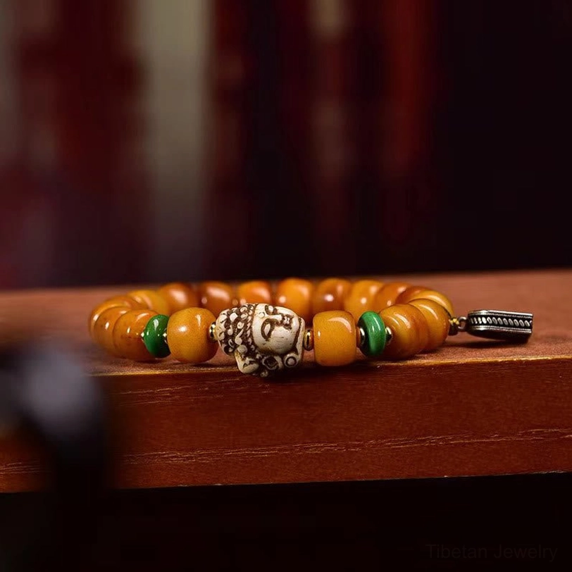 Side view of Tibetan prayer bead bracelet showing amber camel bone beads, green spacers, detailed carved buddha face bead, and metal charm on wooden shelf