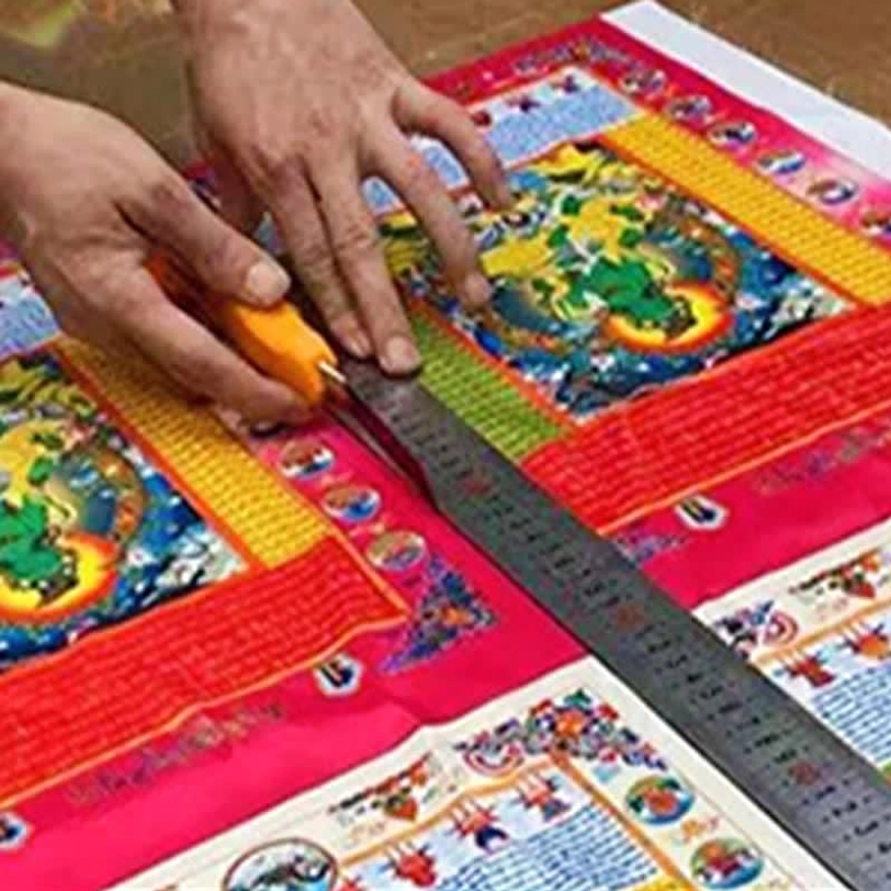 Tibetan prayer flag manufacturing process showing artisan cutting and processing printed flag fabric using ruler and cutter on workshop table