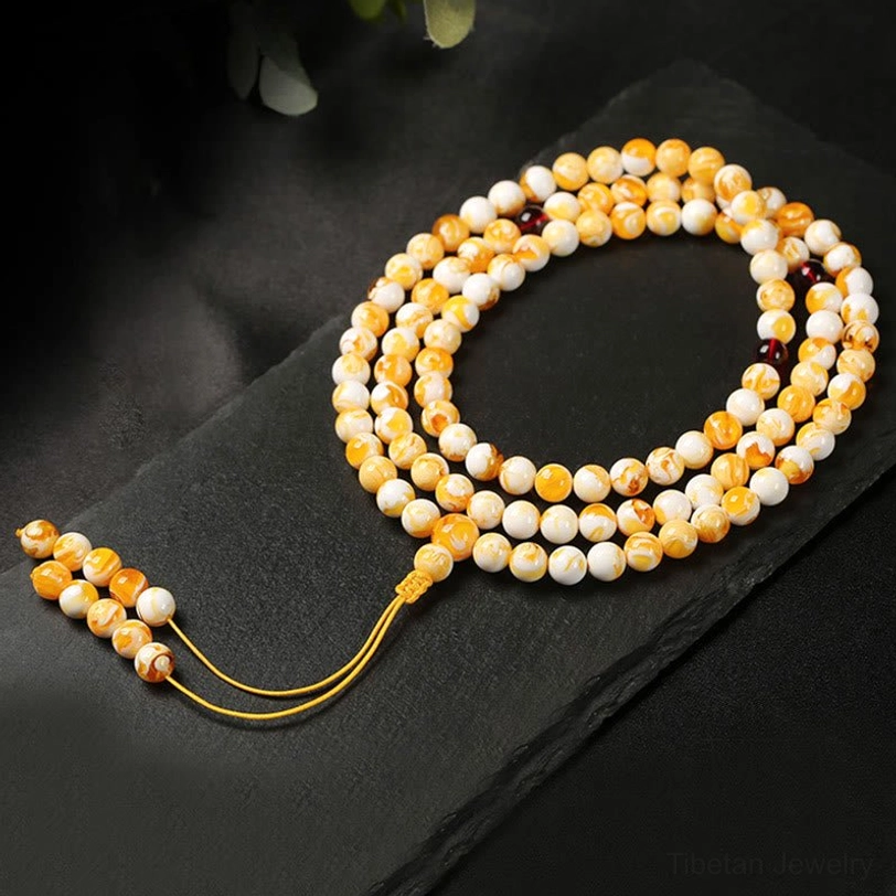 108-bead Tibetan prayer mala with round yellow and white beads, long golden tassel pendant, flat lay on black background