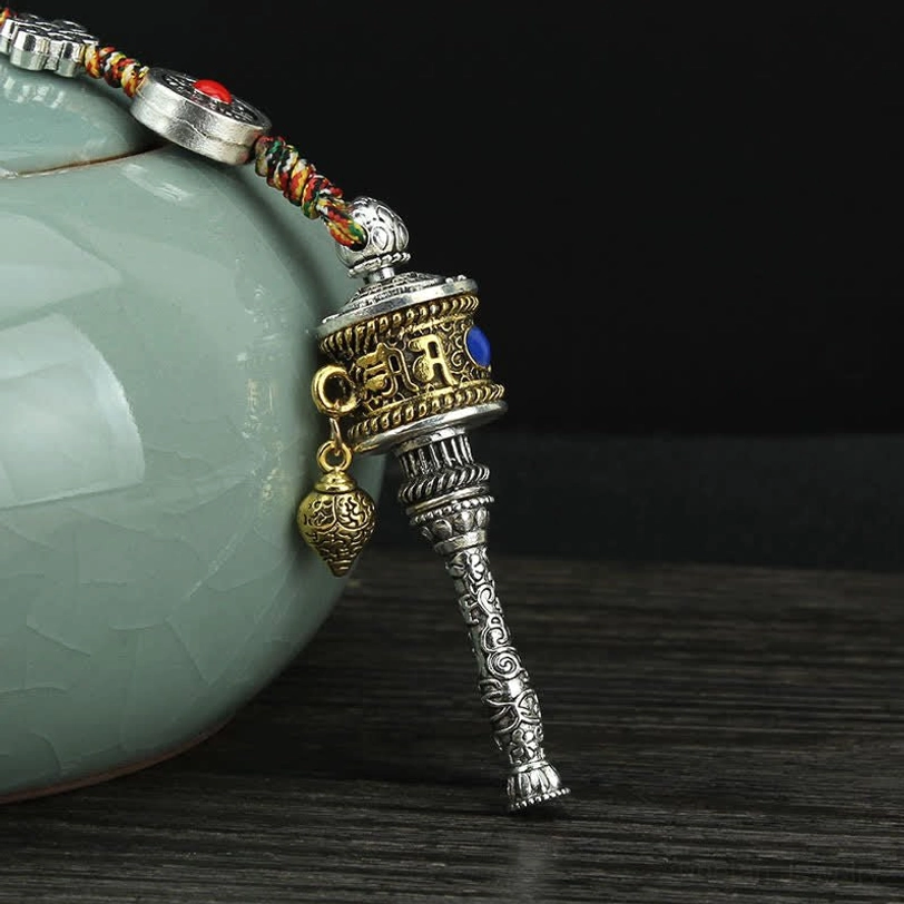 Tibetan prayer wheel hanging accessory with silver body, blue-red gemstone decorations, carved metal handle, and metal bell pendant against celadon jar