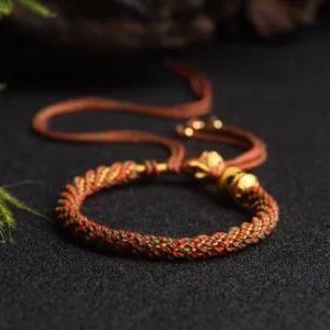 Red-brown handwoven Tibetan prayer wheel lucky bracelet with gold metal accents and adjustable tassel, shown from oblique top view
