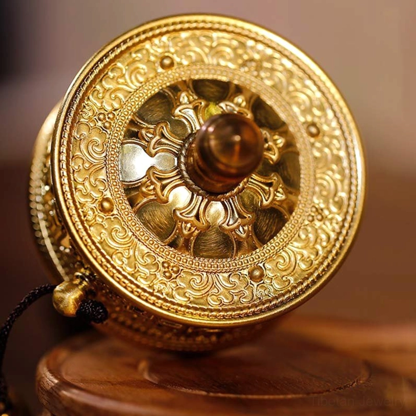 Tibetan prayer wheel front end-face on wooden display stand, featuring bright gold tone with detailed relief carvings, brown sandalwood knob, and attached hanging strap