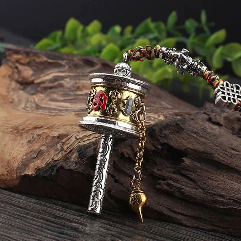 Tibetan car hanging prayer wheel with gold accent on silver body, chain pendant, and colorful braided rope on wooden log display