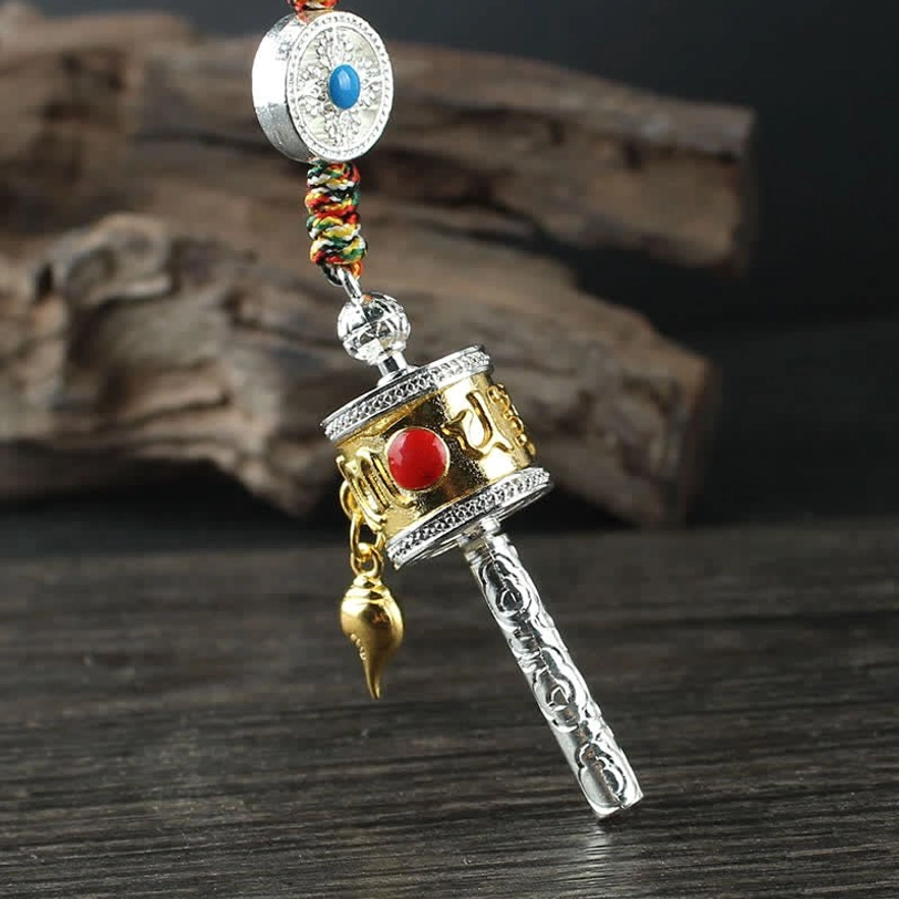 Tibetan prayer wheel hanging decoration with gold-accented body, red gemstone inlay, circular turquoise bead, and braided hanging rope on wooden slab