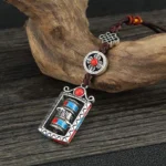 Om Mani Padme Hum prayer wheel keychain with rotatable mechanism, featuring silver alloy, red and blue accents, and dark brown Chinese knot cord on wooden surface