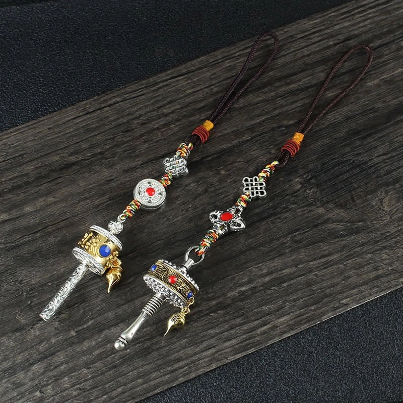 Pair of Tibetan Buddhist prayer wheel pendants with mixed silver and gold tones, brown braided ropes, and decorative prayer beads on dark wooden board