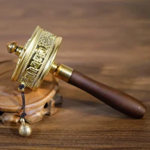 Tibetan prayer wheel with gold-tone metal wheel and dark sandalwood handle, featuring Sanskrit mantra carvings and brass bell pendant on wooden display stand