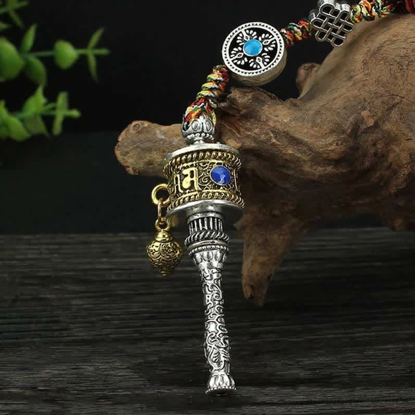 Tibetan prayer wheel hanging decoration with silver tone, blue gemstone inlay, colorful braided rope, and metal drop pendant on wooden root display