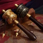 Two handcrafted tibetan copper prayer wheels with carved mantras and wooden handles displayed together on wooden table
