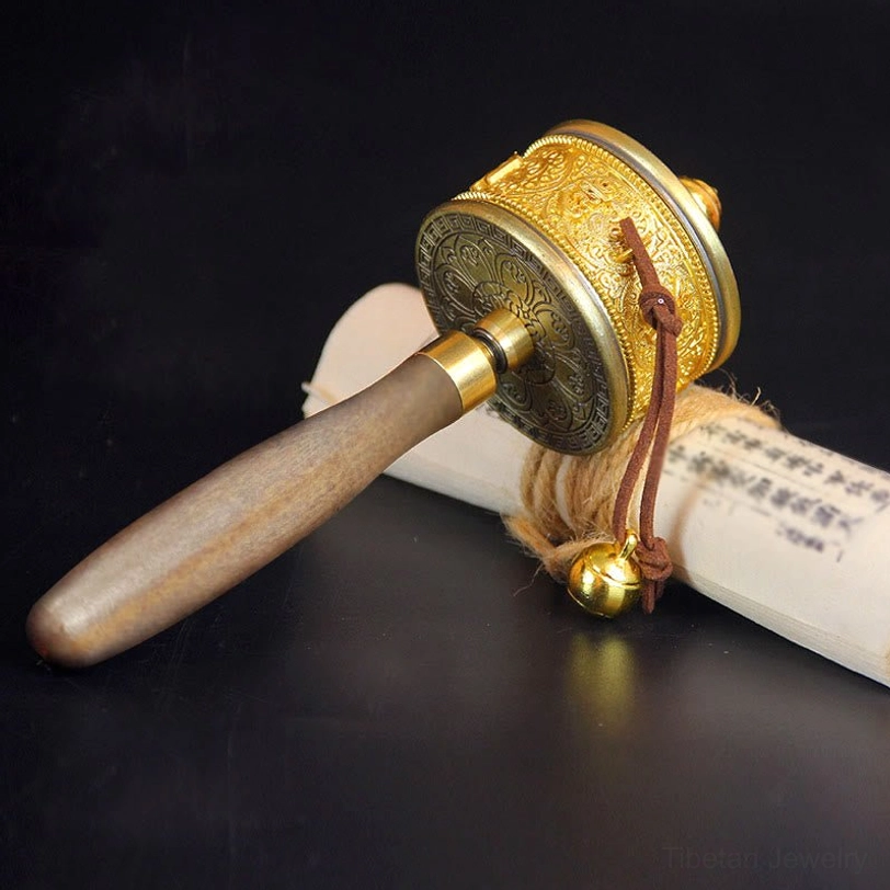 Handheld Tibetan prayer wheel with solid wood handle and brass bell pendant displayed beside rolled Buddhist scroll